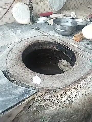 A person is shown preparing naan bread in a tandoor oven. The oven is cylindrical with a circular opening on top, built into a concrete or brick base. The person uses their hands to carefully place the flatbread dough against the inner wall of the oven. Several other unbaked flatbreads are visible on a surface nearby, along with two metal bowls, one containing water, a small plate, and a few other items. A long metal skewer rests beside the oven. The surrounding environment appears to be a kitchen or food preparation area.