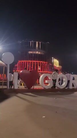 The video shows a nighttime scene with a large, illuminated sign that reads "I LOVE GORAKHPUR". The sign is composed of large, white, three-dimensional letters, with a red heart representing "LOVE". In the background, there is a multi-tiered structure with orange and yellow lights. Several people are visible, some standing near the sign, and others walking past. The ground is paved and appears to be a walkway or public area. Streetlights are also visible, casting light into the scene.