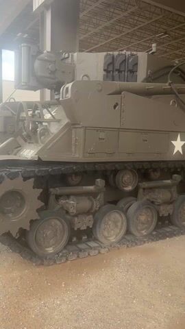 The video shows a large, open-air warehouse or storage facility filled with military vehicles. A tracked vehicle, possibly a tank recovery vehicle or engineering vehicle, is in the foreground, painted in olive drab and marked with a white star and the serial number "USA 30141318."  The camera pans back to reveal a row of similar vehicles, including trucks and other military transport, lined up under the metal roof structure. The ground is covered in a light-colored dirt or gravel. The environment appears dusty, suggesting a dry climate. There is no visible activity, people, or interaction within the space.