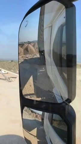 From the vantage of a side-view mirror of a white truck, the camera shows the truck driving past piles of dirt on a dirt road. The sky is clear and blue. The dirt piles are next to a low rock wall, and beyond them, the terrain is flat. A crane is on the road in front of the truck.