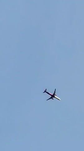 A plane flies across a clear blue sky. The plane is seen from below and is angled slightly to the left. It is a narrow-body jet with white wings and a red stripe along the fuselage. The plane is in the distance and appears to be moving from left to right across the frame.