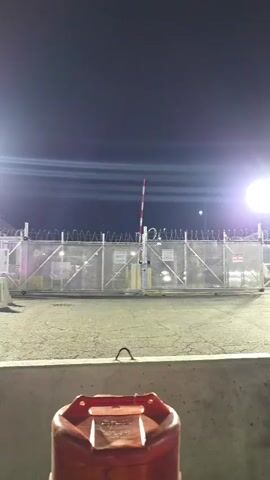 The video presents a nighttime scene, likely at a secure or industrial facility in Newark, United States. In the foreground, a red plastic fuel can with a black handle and cap is visible, placed in front of a concrete barrier. Some text is embossed or printed on the fuel can, but it is too small to read clearly.

Behind the concrete barrier, a prominent chain-link fence forms a security perimeter. This fence is topped with multiple strands of razor wire, also known as concertina wire. Behind the main fence, a security gate system is visible, including a red and white striped barrier arm, typical of controlled access points. More fencing and additional concrete blocks are discernible further back.

The scene is brightly illuminated by several powerful artificial floodlights, creating intense lens flares, particularly a large circular flare on the right side of the frame. These lights cast a strong glow, and some areas behind the fence exhibit a faint purple hue. The background is a dark, clear night sky, with some horizontal light streaks possibly caused by distant lights or camera effects.

Although no individuals are visually present in the frame, the audio features a clear and loud chant of "The people united will never be defeated." This widely recognized protest chant indicates that a demonstration or gathering is taking place at or near this secure location.
