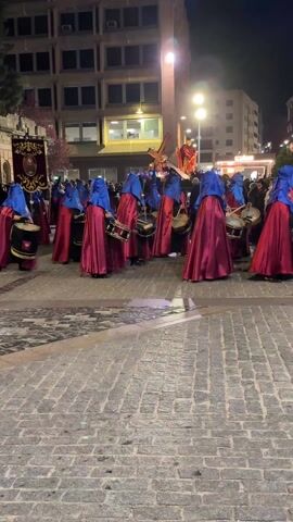 At night, a religious procession is taking place in a city square. The ground is paved with gray cobblestones. At least a dozen people, dressed in hooded red robes and blue hoods, are playing drums. They are arranged in a rough circle. Behind them, a religious banner can be seen, along with a statue of a robed figure holding a cross. A crowd of onlookers watches the procession. Multi-story buildings and streetlights can be seen in the background.