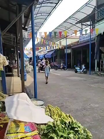 The video shows a street market. In the foreground, there are piles of produce, including corn and leafy green vegetables, along with some packaged goods. A young child, dressed in light blue shorts and a t-shirt, stands in the middle of the street, holding something to their mouth and looking towards the camera. The child is standing between two blue support poles. In the background, other people are visible, some riding motorbikes and others walking. The street is covered by a translucent awning supported by poles, and colorful triangular flags are strung across the width of the market. Buildings line the sides of the street, appearing to be shops or stalls. The overall impression is of a busy, open-air market on a sunny day.