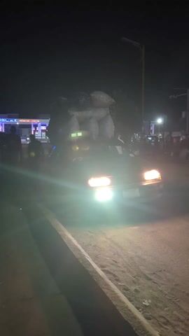 At night, a car overloaded with large sacks is parked near a curb, with the car's headlights on. People mill around the car and a nearby Shell gas station with its lights illuminated. A stack of rectangular blocks is positioned between the camera and the car. A man in a red shirt walks past the blocks. Someone holds a phone to record the scene.