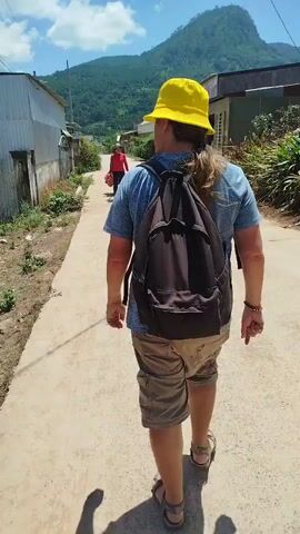 A person wearing a yellow bucket hat, blue t-shirt, and khaki shorts walks away from the camera down a paved path. They have a black backpack on and are wearing sandals. Another person wearing a red shirt and a hat is further down the path, pulling a red cart with two large red objects in it. The path is lined with simple buildings and some vegetation. In the background, there are mountains under a bright, cloudy sky.
