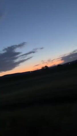 The video shows a car driving along a dirt road at dusk. The sky is a deep blue with wisps of clouds, and a faint orange glow can be seen on the horizon, likely from the setting sun. The landscape is largely dark, with rolling hills in the distance. The headlights of the car illuminate the road ahead, which is flanked by tall grass and vegetation. The road appears unpaved, and the car seems to be moving at a moderate pace.