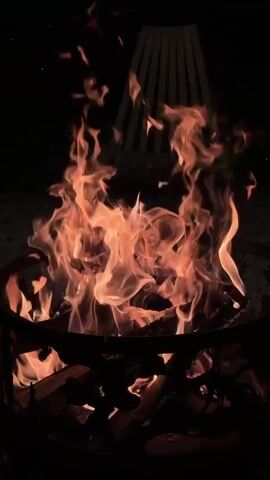 A close-up, vertical shot captures a roaring campfire in the dark. Flames of orange and yellow dance and flicker upwards, consuming logs within a dark metal fire pit. The fire pit has decorative cutouts. In the background, beyond the fire, a dark, indistinct object with vertical lines is visible, possibly a chair or fence. The overall environment appears to be outdoors at night.