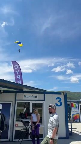 The video captures a scene at a skydiving center on a sunny day. A person is descending with a yellow and blue parachute against a bright blue sky dotted with white clouds.  In the foreground, a white, modular building labeled "Manifest" is visible. A woman in purple overalls stands near the entrance, and a man with a cap and beard stands beside her. The building has multiple windows, and an umbrella with the words "Rigging Loft" is visible near a separate unit. The number "3" is painted on the side of the building.  Flags are flying on the right, and mountains are in the background.