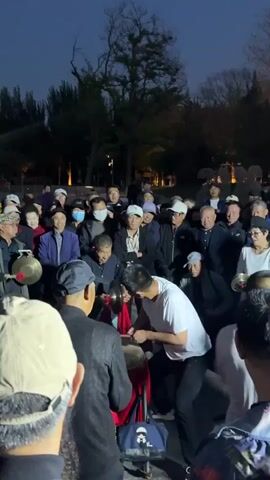 A group of people, mostly elderly and wearing caps, are gathered in an outdoor setting, possibly a park. Some of them are wearing face masks. In the center of the crowd, a younger man wearing a white t-shirt is energetically playing a red drum, while another man next to him plays cymbals. Both of them are smiling and animated. The surrounding environment consists of trees and what appears to be park lighting. The scene is captured with a handheld camera, placing the viewer within the crowd. The overall atmosphere suggests a festive or celebratory occasion.
