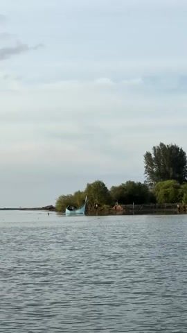A light blue boat is docked near a lush green island. The boat has a raised stern and a pointed bow. The island is covered in trees and foliage, with a few darker trees forming a dense patch on the right side of the island. The water in the foreground is calm and reflects the overcast sky. The sky is a pale blue with scattered clouds. The overall scene is serene and natural.