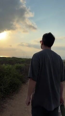 The video shows a person walking away from the camera on a dirt path towards the ocean. The person is wearing a dark t-shirt and shorts. They are carrying a water bottle in their right hand. The sun is setting, casting a warm glow on the scene. There are bushes and vegetation on either side of the path, and the ocean is visible in the distance. The sky is cloudy with patches of blue.