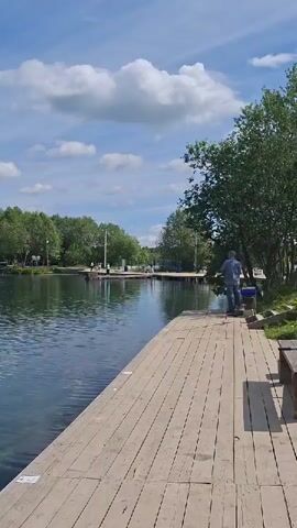 The video captures a serene outdoor scene on a bright, sunny day in Kipen, Russia. The primary setting is a wide, calm body of water, likely a lake or a wide river, with its surface reflecting the clear blue sky and fluffy white clouds.

In the foreground, a long, wooden pier or dock extends along the right side of the frame, providing access to the water. The pier is made of light-colored wooden planks. On this pier, a man dressed in a blue shirt and dark trousers is visible, standing near the edge of the water. He appears to be engaged in an activity, possibly fishing, as there's some equipment next to him, although the details are not clearly discernible.

Across the water, on the opposite bank, a line of lush green trees forms a natural backdrop. Further down the water, in the distance, more people can be seen on what looks like another section of the pier or a distant bank, suggesting a popular recreational area. The trees also line the bank on the right side, partially obscuring the view beyond the pier.

The sky is a vibrant blue, dotted with scattered, well-defined white clouds. The overall impression is one of peace and tranquility, characteristic of a pleasant day spent outdoors by the water. There is no visible text in the video.