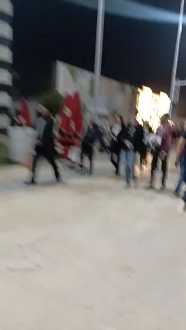 The video shows a group of people walking in what appears to be a plaza at night. Several are wearing jackets or suits. The plaza has red picnic tables with umbrellas, and there are buildings visible in the background. People are moving in various directions.