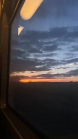The video shows a view from a train window at sunset or sunrise. The sky is a mix of dark gray clouds and a bright orange glow on the horizon. The silhouette of the landscape is visible in the foreground. There is a light fixture visible in the interior of the train above the window.