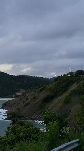 This media file captures a scenic view of a coastal landscape, likely in Rawai, Thailand. The scene is dominated by a lush, green hillside that slopes down towards the turquoise ocean. Waves crash against the rocky shore, adding a dynamic element to the otherwise tranquil setting.

At the top of the hill, a white windmill stands prominently against a cloudy sky, suggesting a source of renewable energy. A small building or structure is visible near the windmill, hinting at human presence in this natural environment. Further in the distance, a paraglider can be seen soaring through the air, adding a touch of adventure and movement to the frame.

The time of day appears to be late afternoon or early evening, as indicated by the muted light and overcast sky. The weather seems to be somewhat cloudy, which can contribute to a serene and perhaps slightly dramatic atmosphere. The presence of palm trees and dense vegetation further reinforces the tropical location.

The overall emotion evoked by the image is one of peacefulness and natural beauty, with a hint of human activity blending into the environment. There is no visible text in the media file.