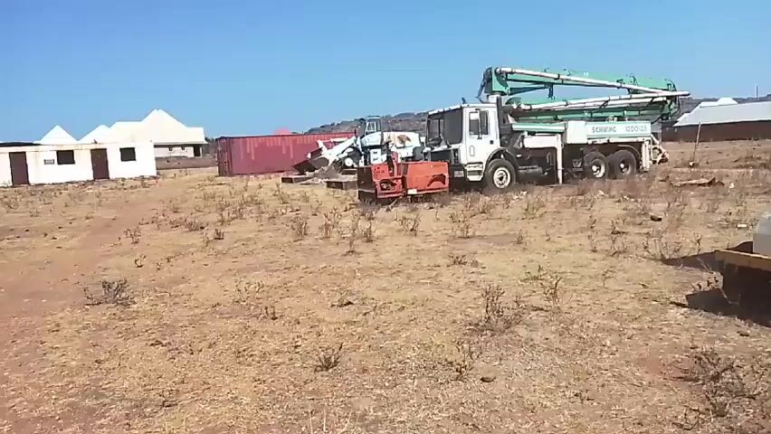 The video shows a construction site in an arid, rural area under a clear blue sky.  Several pieces of heavy machinery are present, including a large white truck with a green concrete boom extending from its back, labeled "SCHWING 1200.23". An orange concrete mixer is parked in front of the truck.  Behind the truck, there's a white front-end loader with a dirt pile in its bucket.  A red shipping container is visible, as well as a small white building with a pyramidal roof and two small buildings with flat roofs, suggestive of residential or storage structures.  The ground is dry and covered with sparse vegetation. The camera moves slightly, showing more of the field.