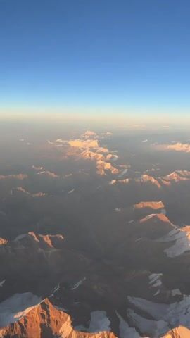 This video provides an aerial view of a vast mountain range bathed in the warm glow of sunset. The sky transitions from a deep blue at the top of the frame to a hazy orange and yellow horizon, indicating the sun is low in the sky. Snow-capped peaks are visible throughout the landscape, with some of the closer mountains illuminated by the golden sunlight, highlighting their rugged textures. Clouds are scattered across the sky, some appearing as wisps near the horizon and others casting shadows on the mountain slopes. The perspective suggests the viewpoint is from an aircraft, looking down and slightly forward. The image is focused on the natural beauty of the mountains and the atmospheric lighting.
