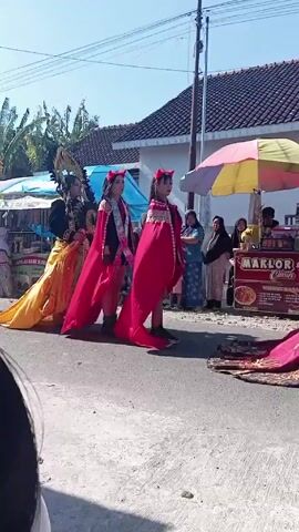 The video captures a street scene in Indonesia, featuring a procession of performers in elaborate costumes. The procession, likely part of a local celebration, is walking along a street lined with houses and small businesses.

The performers wear various costumes, one notable figure has a long golden gown and a headdress. Two others wear red capes and devil horns. Spectators, including women in traditional attire and children, are watching the procession. A vendor's stall selling snacks is visible in the background.

The weather is sunny, and the environment suggests a rural or suburban area in Indonesia. The camera viewpoint appears to be from the side of the road, providing a clear view of the procession and the onlookers.