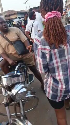 The video shows a street scene in what appears to be a West African city. The camera follows a young person with brown, braided hair and a pink hair tie. They are wearing a plaid shirt and dark shorts. The street is crowded with people, motorcycles, and a yellow auto-rickshaw. Buildings line the street, some painted in bright colors. The scene appears bustling and lively.