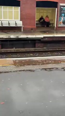 The video shows an outdoor scene at a train station. Two individuals are sitting on a bench within a sheltered waiting area constructed of red brick. The platform is wet, with leaves scattered across the surface. Train tracks run parallel to the platform. A poster is affixed to the brick wall near the waiting area. The day appears overcast.