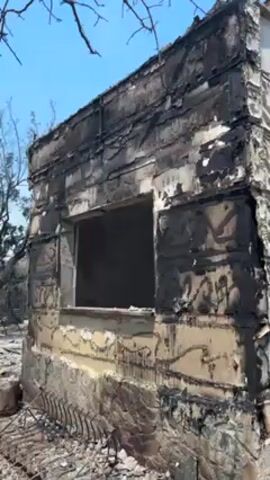 The video shows the exterior of a badly fire-damaged building on a bright, sunny day. The structure appears to have been a two-story building, but the upper story is heavily damaged with only the bare structural elements remaining. The exterior walls are charred, peeling, and have black soot streaks, while a window opening has no glass and only the frame remaining. A ladder leans against the side of the building, and a section of metal fencing lies on the ground in front. The surrounding ground is covered with debris and ash. Smoke or steam can be seen rising from within the building's remains. Bare tree branches are visible in the upper corners of the frame, and an olive tree can be seen in the background.