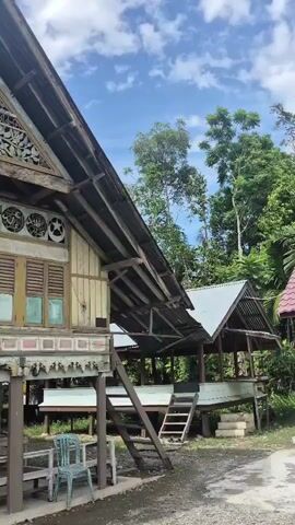 A low-angle, medium shot shows an ornate, weathered wooden building with a steep roof, intricate carvings on its gable, and multiple louvered windows. The building is elevated on wooden stilts, revealing a concrete area underneath with a plastic chair and a table. A small, lighter-colored outbuilding with a similar roofline is visible to the right, also elevated on stilts with a wooden ramp leading up to it. Lush green trees form the background against a bright, cloudy sky.