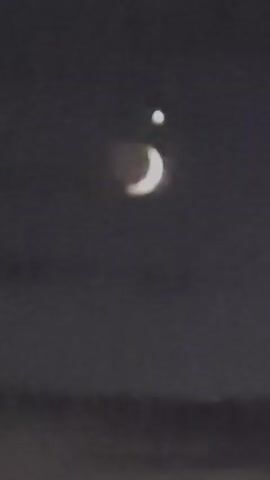 A crescent moon and a bright star are visible in the night sky. The camera pans across the sky, showing the moon and star in different positions. The video then shifts to a view of a residential area at night, with houses and streetlights visible. The moon and star are still visible in the sky.