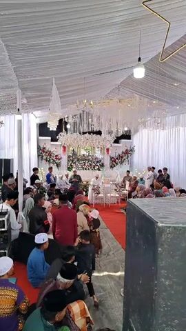 Inside a white tent, a Muslim wedding ceremony is underway. A group of people, mainly men and children, are seated on the floor, facing a stage area. The stage features white chairs for the wedding couple, flanked by floral arrangements and white curtains. The tent is decorated with chandeliers and lights. A large speaker is positioned in the foreground. The people are dressed in a mix of traditional and casual attire, many wearing Muslim caps. The atmosphere is formal and respectful.