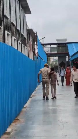 A police officer in uniform walks with a young person on a tiled walkway. The officer has his arm around the young person's shoulder. They are walking away from the camera, towards a covered walkway with blue roofing. To their left is a tall, corrugated blue metal fence, behind which is a building with white and black siding and windows. To the right of the walkway, other people are visible, some walking away from the camera. The sky is overcast.