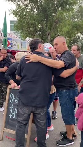 Here's a detailed description of the video:

The video shows a group of people in an outdoor setting. A man in a black shirt and jeans is embracing another man in a black shirt and denim shorts. They appear to be having an emotional exchange, possibly a warm greeting. To the left, a cameraman is filming them with a professional video camera. Other people are standing around, some observing the interaction. A small blackboard is positioned in front, displaying "Shawarma $15.00" written in chalk. In the background, there are food stalls and tents, some with Cuban flags and "Cuba" written on them. Trees are also visible in the background. The overall atmosphere seems to be that of a festival or public gathering.