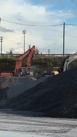 An orange excavator is shown on the left, and a black excavator is on the right. There are large piles of gravel in the foreground and middle ground. In the background, there are power lines and poles. The sky is cloudy. The orange excavator is digging into a pile of gravel.