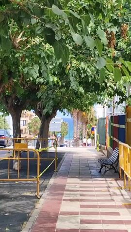 The media file captures a view down a tree-lined sidewalk in El Campello, Spain. The scene is bathed in sunlight, suggesting it is daytime, likely late morning or early afternoon, with a clear sky. Lush green trees overhang the sidewalk, providing shade. The sidewalk itself is paved with a decorative pattern of red and cream tiles.

In the foreground, yellow safety barriers with yellow and black caution tape are visible, indicating some sort of work or closure in the area. A park bench with a striped cushion sits to the right of the sidewalk. Beyond the immediate foreground, the sidewalk slopes gently downhill towards the sea, which is visible in the distance. Palm trees and buildings line the street, with a car parked on the left side.

Further down the street, a few people are visible, casually walking and seated, enjoying the pleasant weather. The overall atmosphere appears relaxed and summery. The presence of the sea and the style of the buildings and palm trees strongly suggest a coastal Mediterranean location, consistent with El Campello, Spain. There are no discernible signs with legible text within the frame.