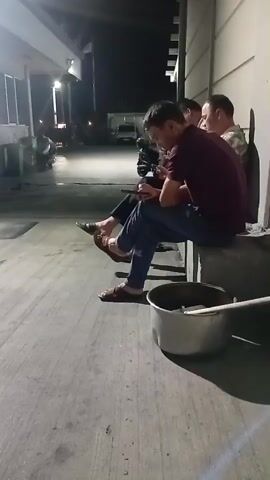 Three men are sitting on a concrete ledge outdoors at night. The man in the foreground is wearing a maroon shirt and blue jeans and is looking at his phone. The man next to him, partially obscured, is wearing a dark shirt and is also looking at his phone. The third man, on the far right, is wearing a camouflage shirt and appears to be looking forward. In front of the men is a large metal pot with a handle. The ground is concrete, and there is a building with lights in the background.