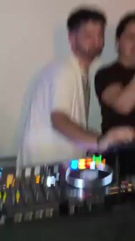 Two people are visible in the video. A man with short dark hair, a beard, and a white t-shirt is in the foreground, operating a DJ mixing console. He is moving his hands quickly over the equipment. Behind him, slightly out of focus, is a woman with dark hair wearing a dark t-shirt. The background is a plain white wall. The video is shaky and the focus is often blurred due to movement. The primary action is the man using the DJ equipment.