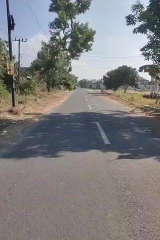A video shows a paved road in what appears to be a rural or suburban area. The road has a white dashed line dividing the lanes. On the left side of the road, there are trees and some vegetation, a utility pole, and a white fence with garbage cans. A person on a bicycle is visible near the garbage cans. On the right side of the road, there are trees, some buildings in the distance, and dirt areas. The sky is clear and blue with some clouds. The camera is moving, giving the impression of a ride down the road.