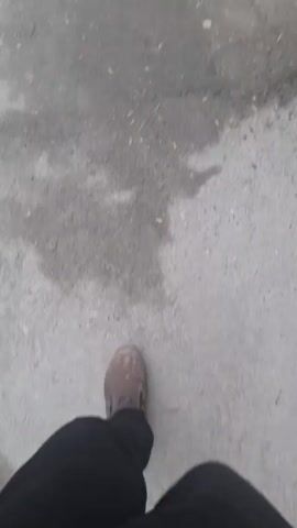 The video shows a person walking along a cracked asphalt street. The street is lined with buildings on either side, one of which is a white building with black tiled trim at the bottom and a gutter. There is a leaning utility pole with a lightning symbol on it on the right side of the street. A person walks away in the distance.