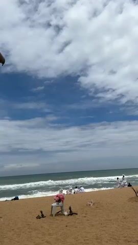 This video shows a beach scene under a cloudy sky. The foreground is a sandy beach with a white plastic chair in the center, adorned with a bouquet of pink flowers. To the left and right of the chair are some small, dark objects that appear to be decorative. Further back on the beach, several people are seated or lying down. Some are wearing white. They are positioned near the water's edge, where waves are breaking on the shore. The ocean is a turquoise green, and the sky is a vibrant blue with scattered white clouds. A dark pole is visible in the upper left corner of the frame, suggesting it is part of a structure or an object above and to the left of the scene.