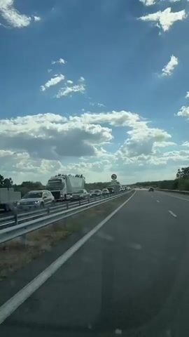 This media file depicts a scene of highway traffic congestion under a bright, cloudy sky. The perspective is from within a vehicle, looking forward along a multi-lane highway. A line of cars and trucks is visible, stretching into the distance, indicating a significant traffic jam.

The setting appears to be a rural or semi-rural area, with trees and greenery visible beyond the highway. The weather is sunny with scattered white clouds, suggesting it is daytime, likely late morning or afternoon. A speed limit sign is visible on the right side of the highway, although the exact speed limit is not discernible. A large white truck with green lettering that appears to say "Vardar Fruit" is prominent in the left lane.

The overall mood conveyed is one of frustration or patience due to the traffic. The image captures a common experience of travel delays on highways. There is no visible indication of people's emotions beyond the implied inconvenience of the traffic. The location context provided is Bozduganovo, Bulgaria.