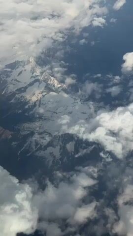 The video shows a view of snow-capped mountains from above, likely from an airplane. Clouds obscure parts of the mountain range, creating a dynamic interplay of white and gray. The sky is visible in the background, ranging from a lighter blue to a deeper, darker blue. The environment is mountainous and appears to be cold due to the presence of snow. No people or actions are directly visible.