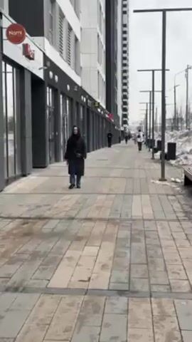 The video depicts an outdoor scene with a tiled pedestrian walkway in an urban area. Several people are walking along the walkway in the same direction, away from the camera. A woman wearing a long, dark coat is walking closer to the camera. Along the left side of the walkway are modern buildings with shops or businesses that appear to be closed. Light poles line the right side of the walkway, with patches of snow visible between the walkway and what appears to be a road. The sky is overcast.