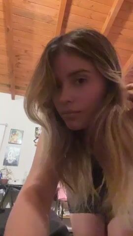 A young woman with medium-length blond hair is standing indoors, looking down at the camera. She has a slight smile and brushes her hair away from her face. The background features a wooden ceiling with beams, a few pieces of art on a white wall, a chair, and a plant. Loud music plays in the background.