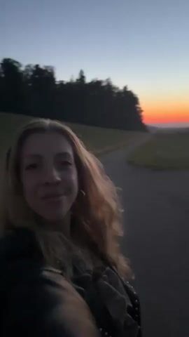 The video begins with a close-up of a woman with blonde hair smiling at the camera. She is wearing a black jacket. The background shows a sunset with an orange and pink sky over a grassy landscape. In the next clip, the woman is still smiling. Behind her, a red car is parked and a person is visible in the driver's seat. The woman is now slightly further away. The following clips show the woman moving, the background becoming blurrier, with lights and possibly buildings visible in the distance. The sky transitions from dusk to a darker blue. The woman continues to smile and move throughout these clips.