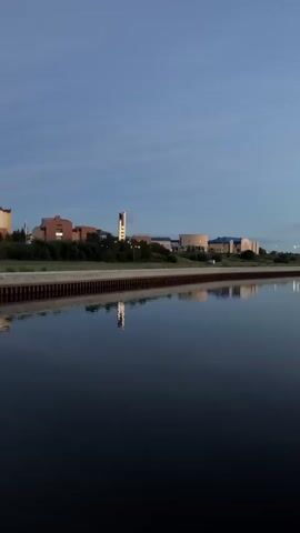 The video shows a calm body of water reflecting the sky and buildings on the opposite bank. The sky is a deep blue with a few wispy clouds. The buildings appear to be modern with some having circular architectural elements. A bridge with a distinctive angled tower is visible on the right side of the frame, also reflected in the water. The scene suggests a twilight or early morning setting, with the lighting indicating a time of day when artificial lights might be on or just turning off. The water's surface is still, creating a near-perfect mirror image of the structures and sky. There are no people visible in the video.
