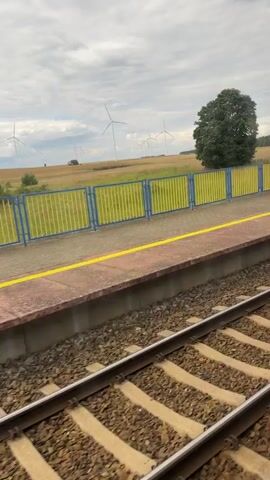 This is a vertical video of a train moving past a rural landscape. The video begins by showing a train station platform with a sign that reads "Burkat." There is a yellow line marking the edge of the platform and a blue fence with vertical bars. In the background, there are fields and several wind turbines visible on a gentle hill. A large, dark green tree stands in the middle ground.

As the video progresses, the view shifts to show more of the train tracks and the passing scenery. The train station shelter with a bench is visible on the left side of the frame. The landscape continues to feature fields, grassy hills, and more wind turbines. The sky is overcast with some lighter patches, suggesting it is daytime, possibly late morning or afternoon. The overall mood is peaceful and observational, capturing a typical journey through the Polish countryside. The presence of the train tracks and the station implies a travel or transit context.
