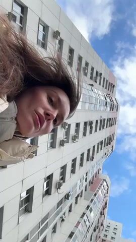 A low-angle, tilted video shows a young woman from a worm's-eye perspective against a backdrop of a tall building and a cloudy blue sky. Her light brown hair is gently swaying around her face, suggesting a light breeze. She is wearing a light-colored puffer jacket over a grey hooded sweatshirt. Her gaze is directed downwards and to the side, giving her a pensive or melancholic expression.

Behind her, a modern, multi-story building dominates the background, featuring numerous windows and external air conditioning units. The building's facade is predominantly white with subtle pink or light red accents. The sky is bright blue with scattered white clouds, indicating daytime. The scene appears to be set in an urban environment, specifically in Edem, Russia. No visible text is present in the video.