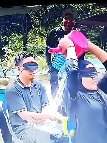 The video shows a group of people participating in a water-based game. In the foreground, a man is seated with his eyes blindfolded, wearing a dark polo shirt. To his right, a person wearing a black headscarf and a dark top is holding a pink bucket and appears to be pouring water. Behind the seated man, another person wearing a black t-shirt with blue stripes is smiling and holding a pink object, possibly a container. Further back, two women are visible, both smiling and looking towards the front. One woman is wearing a red t-shirt and the other a blue headscarf. The environment appears to be outdoors, possibly in a recreational area, with trees and some sort of structure in the background. Water is visibly splashing.
