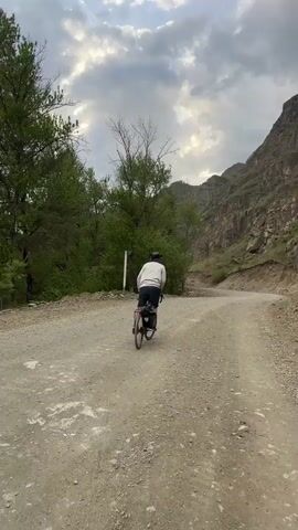 A man is riding a bicycle on a gravel road. The road is winding through a mountain pass. The man is wearing a light-colored jacket, dark shorts, and a hat. The bicycle has panniers attached to the back. The sky is cloudy. Trees are visible on the left side of the road, and a rocky mountain is on the right side.