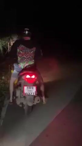 Two people are riding motorbikes on a road at night. Both drivers are wearing helmets and the motorbikes have their headlights and tail lights on. The road appears to be asphalt. Vegetation is visible on the side of the road.