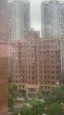 Here is a concise description of the video:

The video displays an exterior view of a cityscape on a rainy day. The perspective is from an elevated position, looking out through a window with raindrops visible on the glass. In the foreground, there is a medium-rise apartment building constructed of red brick with multiple windows and air conditioning units. Beyond this building, several high-rise apartment towers dominate the background, appearing somewhat hazy due to the weather. The buildings are surrounded by green trees and a winding road, adding to the urban landscape.  The scene has an overall tone of a quiet, overcast day in a densely populated city.